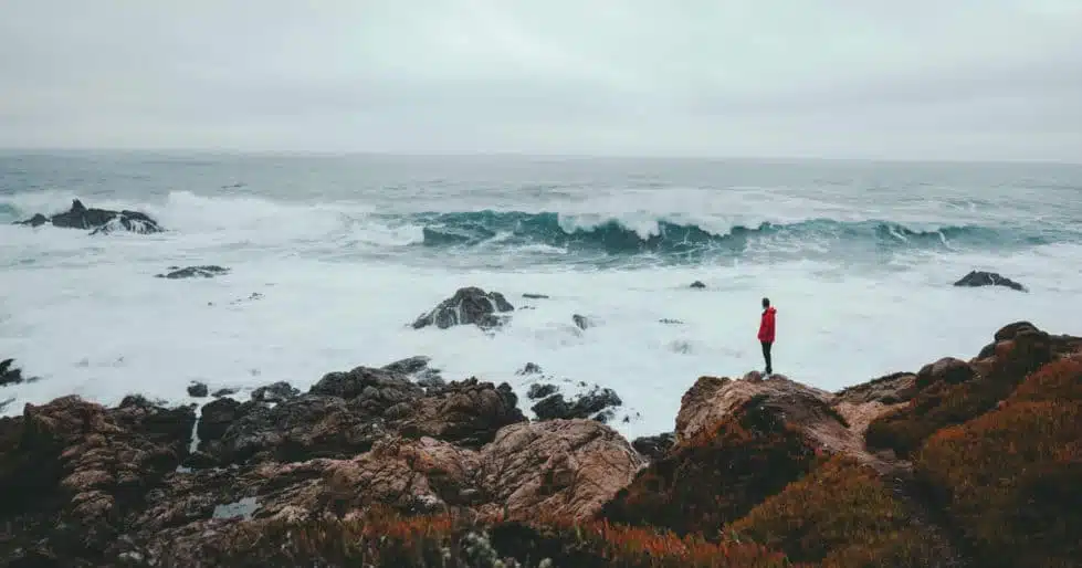 A man in a red coat stands on a rocky shoreline and looks out at the turbulent, white water rolling in.