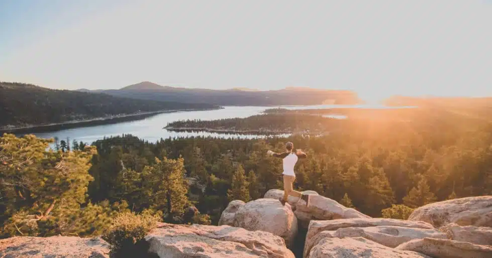 A man, atop a rocky summit overlooking a vast forest and river, leaps from one rock to another.