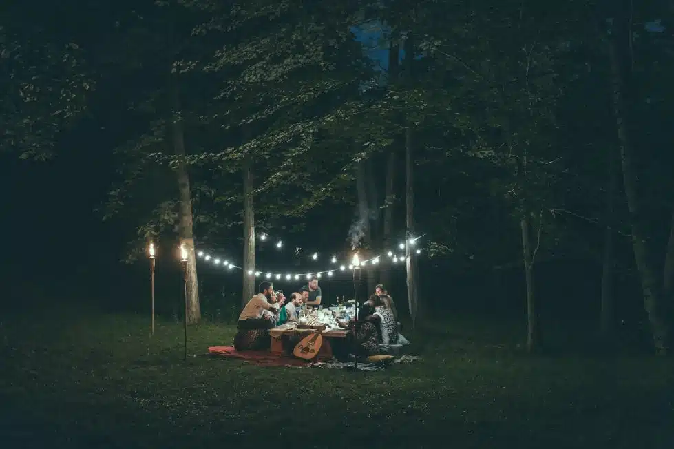 A group of people in a forest at night around a picnic table illuminated by white lights